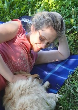 Picture of a woman lying in the grass and petting a dog, which is sitting next to her